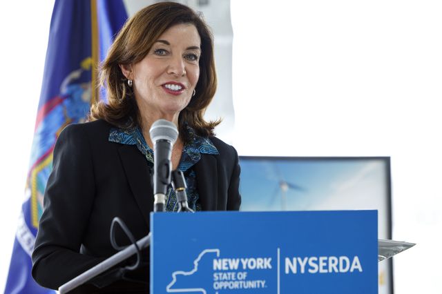 Kathy Hochul, in a black jacket and blue patterned shirt, standing behind a lectern